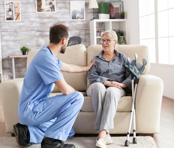 Male health visitor in a nursing home talking with retired old woman while sitting on couch. Old woman with crutches.